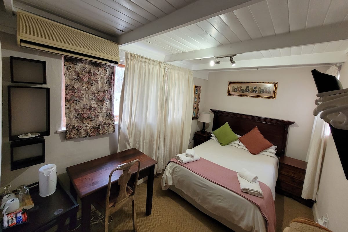 Habitación de hotel con techo de madera, cama con almohadas, escritorio con silla, mesita de noche con lámpara y ventana con cortinas.