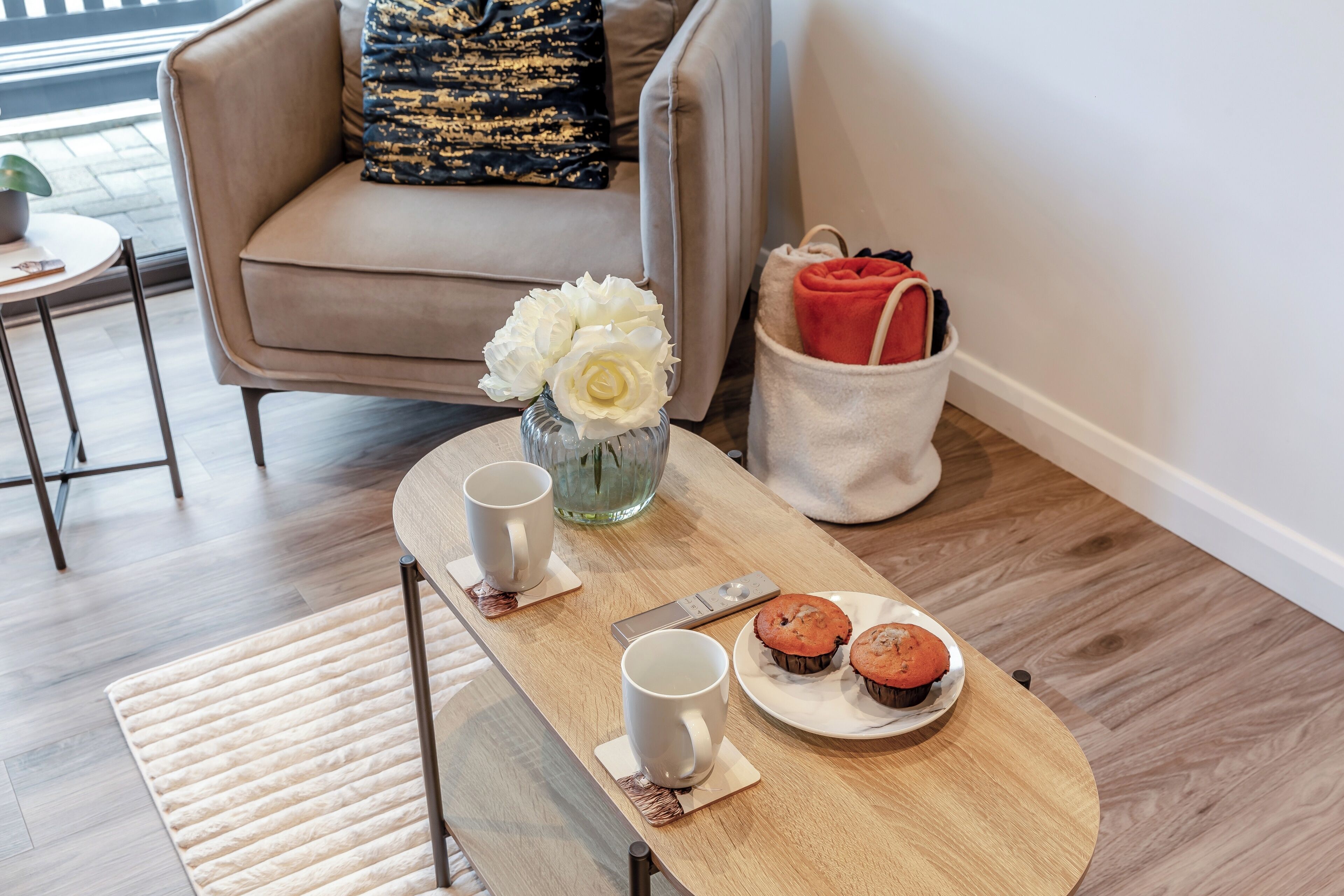 A wooden table with two white mugs, a plate with two muffins, and a vase with white flowers.
