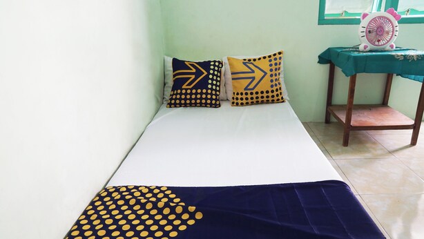 A bed with a navy blue and yellow polka dot blanket, two pillows with arrow patterns, and a small table with a fan on a teal tablecloth. - Hotel O Wunut Homestay Syariah (Sidoarjo)
