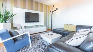 A modern living room with a flat-screen TV mounted on a white wall, a sofa, a coffee table, and a floor lamp. - The View by Atlantic Holiday (Funchal)