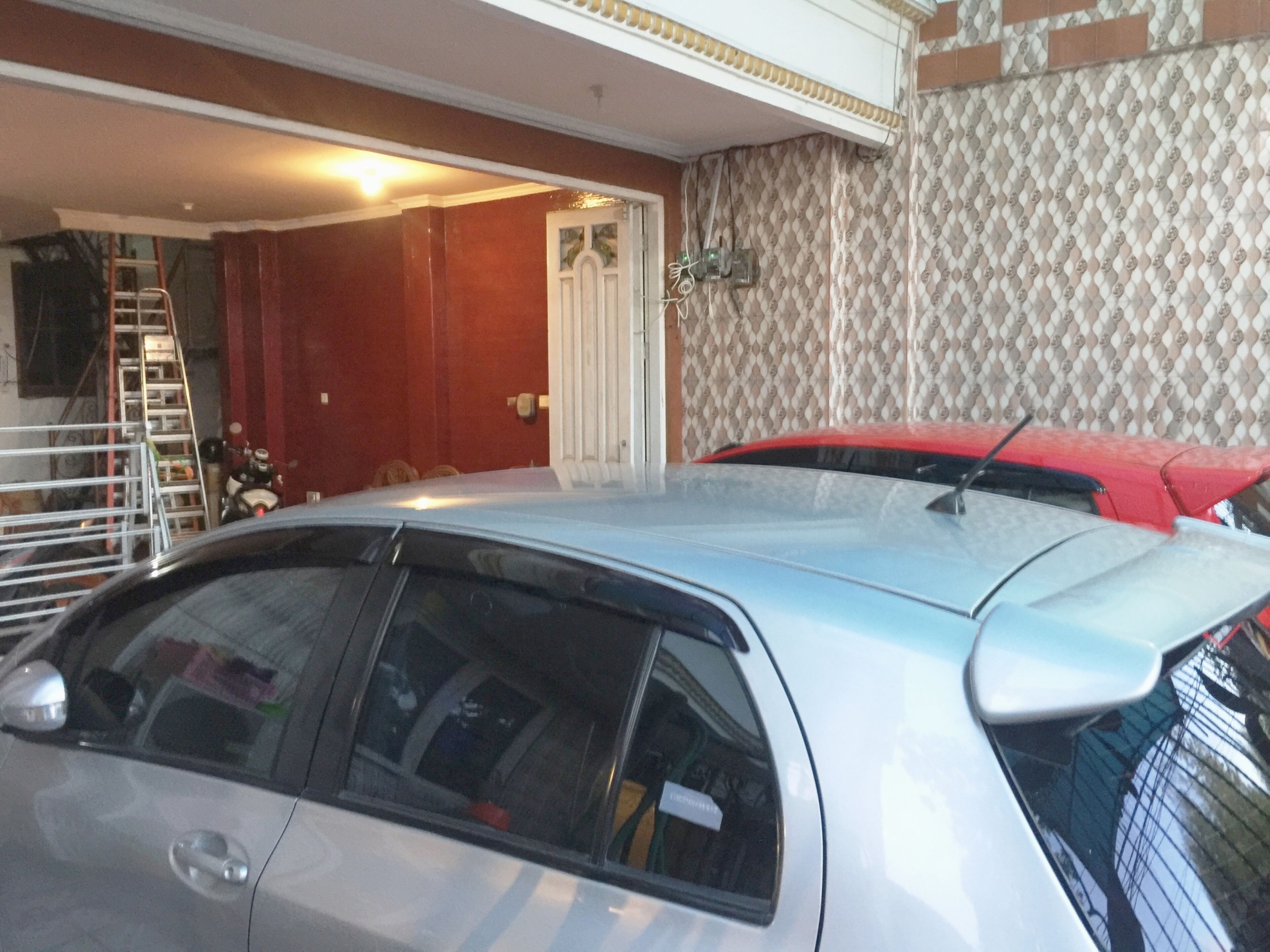 Een garage met een witte auto en een rode auto erin geparkeerd. Een ladder staat tegen de muur en er is een deur naar een andere ruimte.