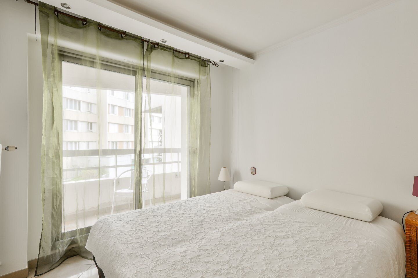 Een slaapkamer met een groot bed, een raam met dunne gordijnen, een nachtkastje en een wandlamp.
