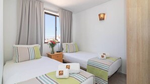 A hotel room with two beds, a window with curtains, a bedside table with a vase of flowers, and a wall-mounted light fixture. - Vilamoura "charming Algardia" T1 (Quarteira)