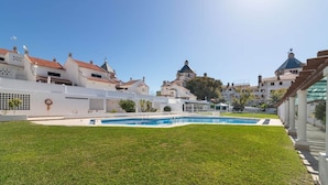 A swimming pool surrounded by a well-maintained lawn, with buildings in the background. - Vilamoura "charming Algardia" T1 (Quarteira)