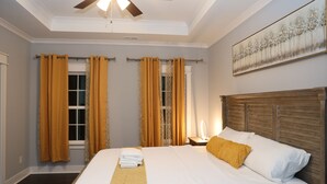 A hotel room with a bed, curtains, a ceiling fan, a painting, and a nightstand with a lamp.