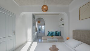 A modern bedroom with a large bed, a sofa with striped pillows, a small table, and a hanging light fixture. - Nevma Suites (Santorini)