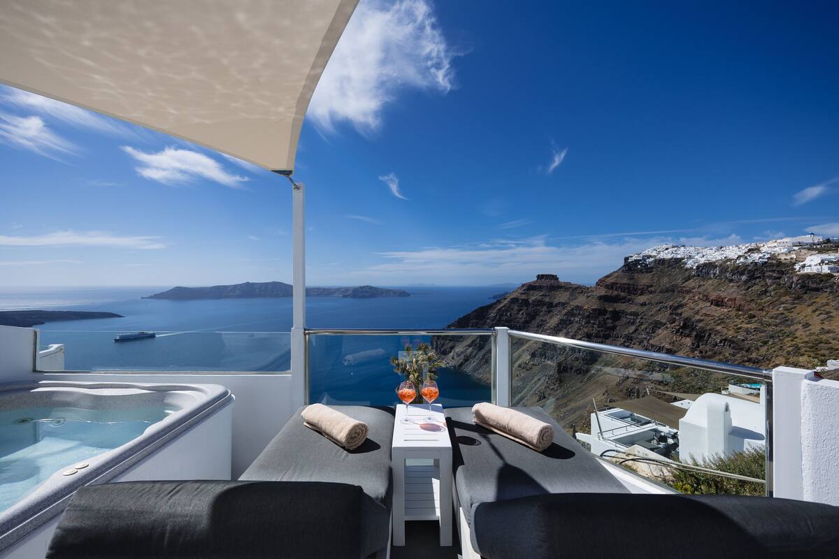 A balcony with a view of the sea and islands, a hot tub, and two lounge chairs with towels.