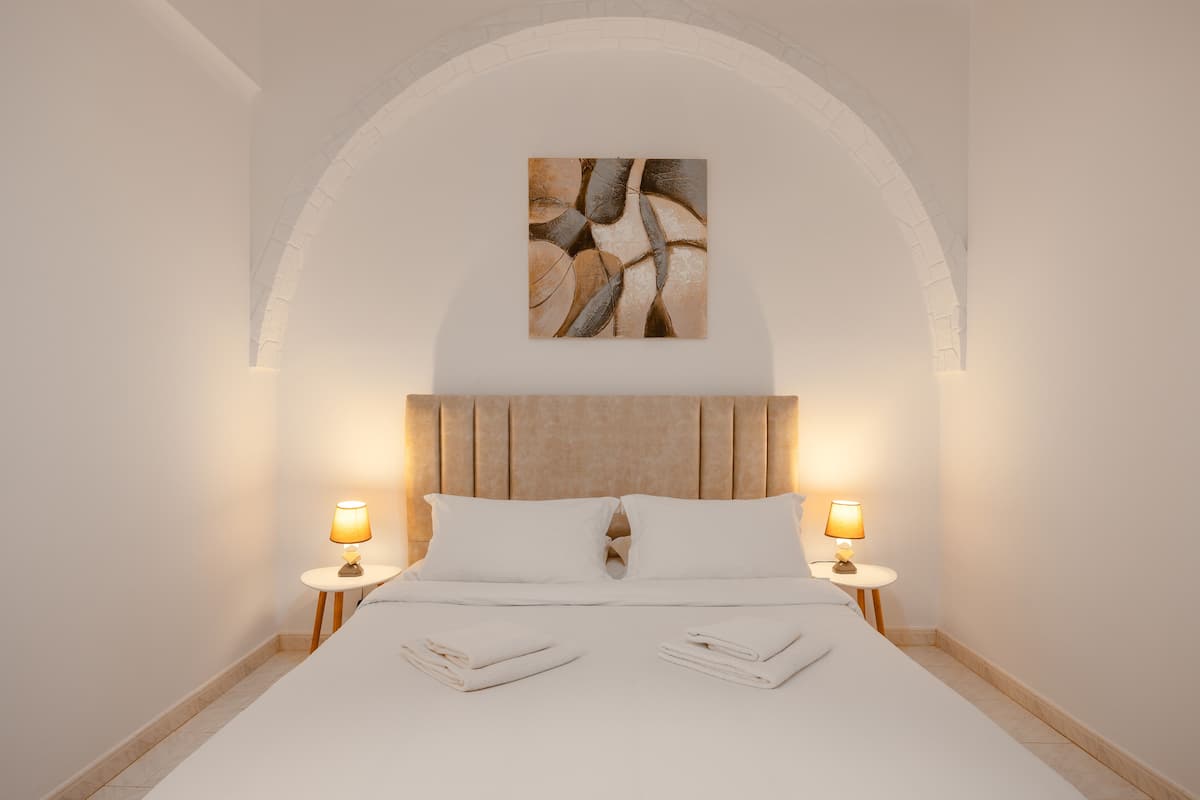 A neatly made bed with white linens and pillows, two bedside lamps, and a painting above the headboard.