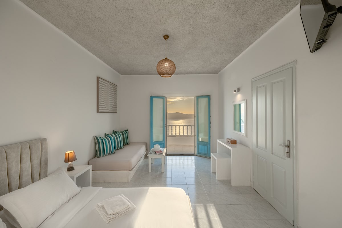 A modern hotel room with a bed, a nightstand, a sofa, a small table, and a view of the outside.