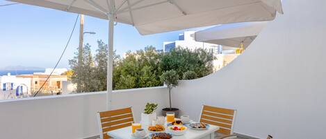 Une terrasse avec une table blanche dressée pour le petit-déjeuner, entourée de chaises en bois, sous un grand parasol.