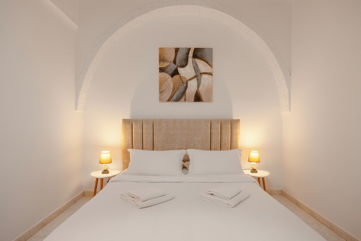 A neatly made bed with white linens and pillows, two bedside lamps, and a painting above the headboard.