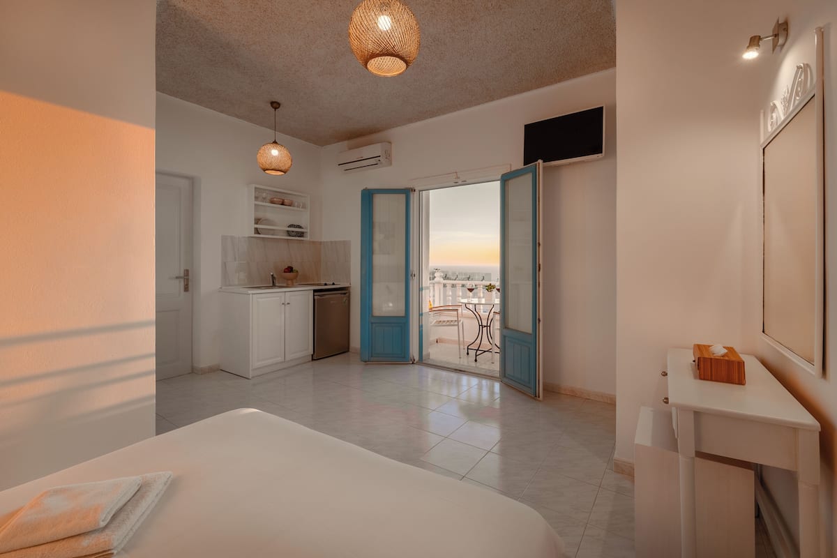 A modern hotel room with a bed, a small kitchenette, a balcony with a city view, and a large window.