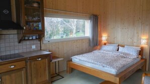 A wooden cabin-style room with a bed, a small kitchen area, and a window with a view of trees. - Studio on Monte Generoso (Castel San Pietro)