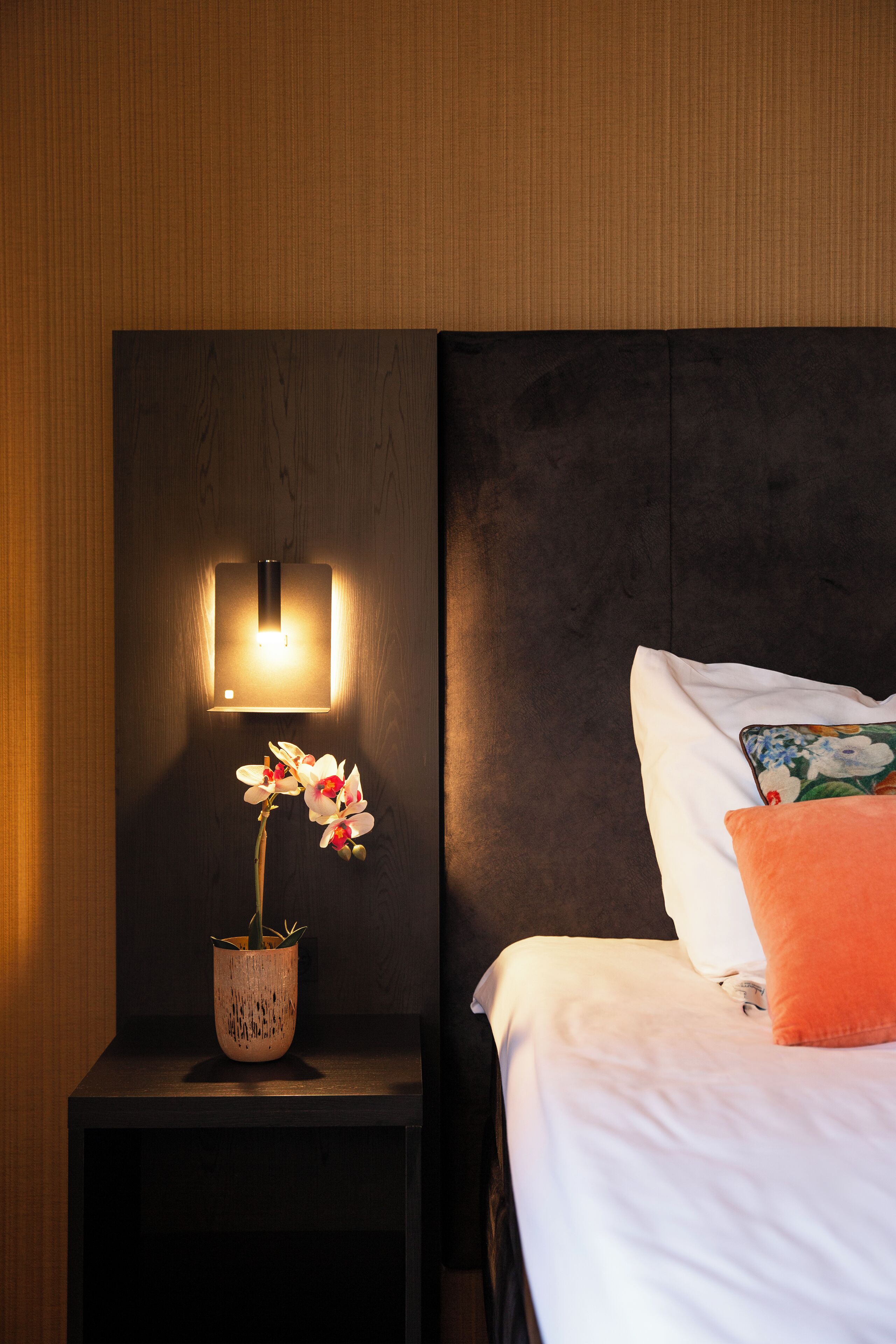 A neatly made bed with white linens and a single orange pillow, a bedside table with a potted orchid, and a wall-mounted light fixture.