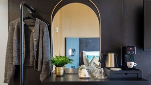 Hotellihuone, jossa on tumma puinen työpöytä, seinälle ripustettu takki, ruukussa oleva kasvi, kahvinkeitin ja peili, joka heijastaa huonetta.