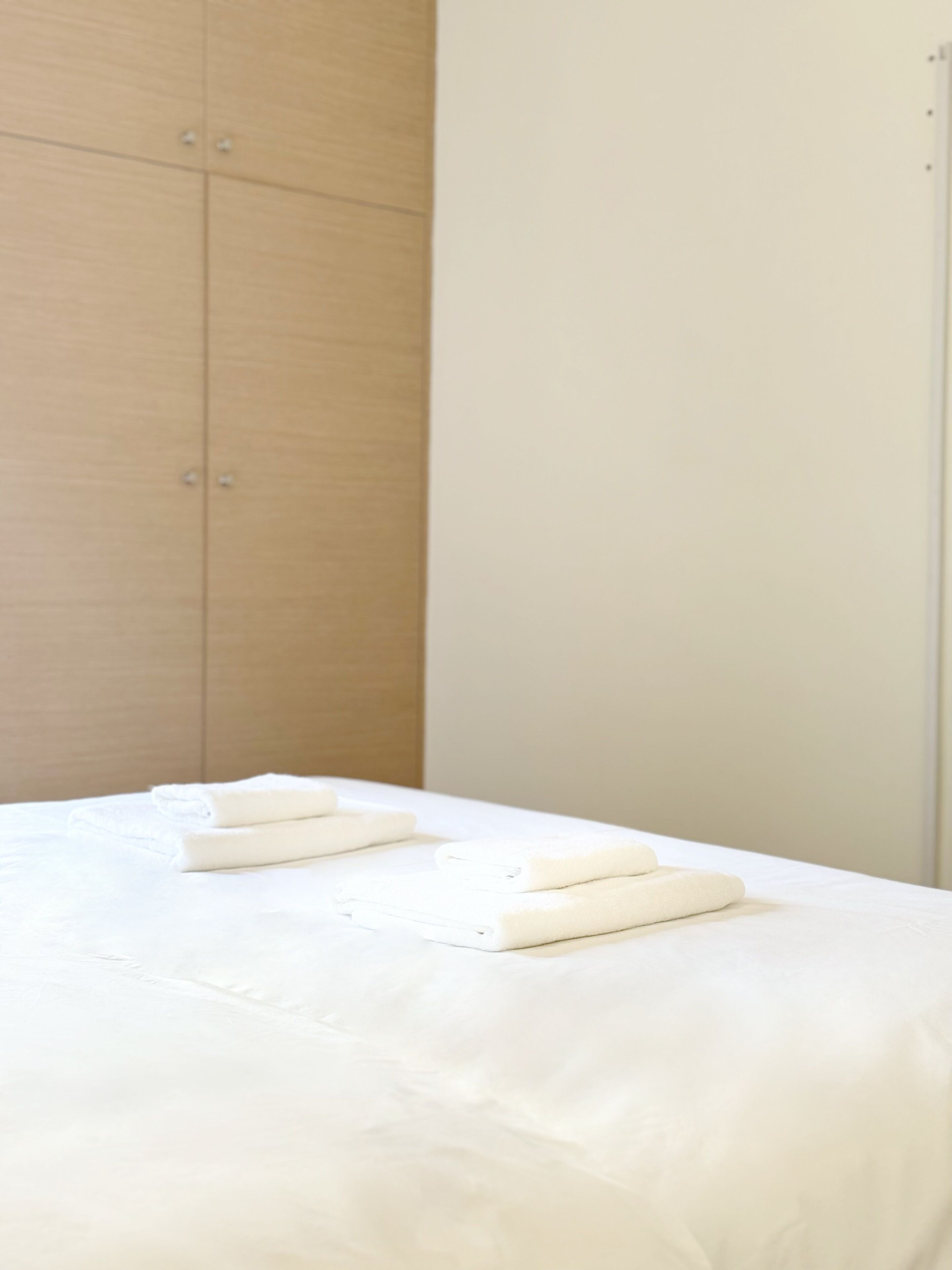 A neatly made bed with white linens in a room with a wooden wardrobe and a plain wall.