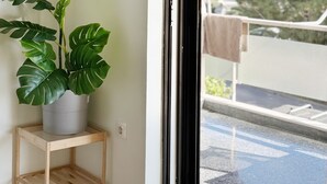 A potted monstera plant on a wooden side table next to a sliding glass door leading to a balcony with a view of buildings and trees. - John ,Home (Glifada)