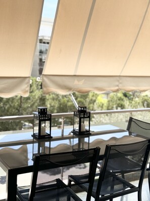 A glass-top table with black mesh chairs, beige canopies, and a view of greenery. - John ,Home (Glifada)