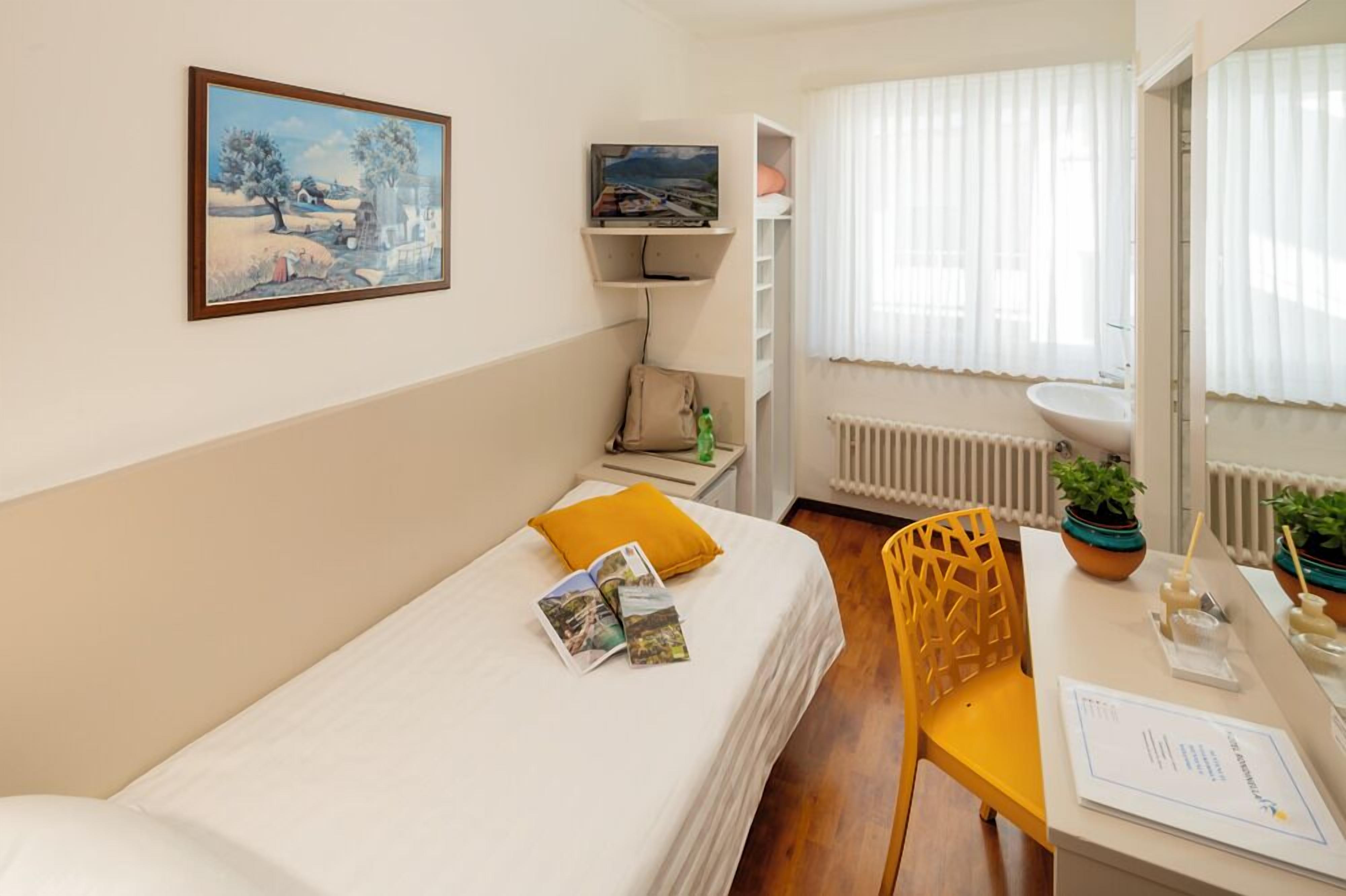 A bedroom with a bed, a chair, a desk, a radiator, a window with curtains, a shelf with a TV, and a painting on the wall.