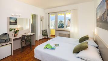 Habitación de hotel con cama, escritorio, silla y ventana con vistas a edificios y montañas.