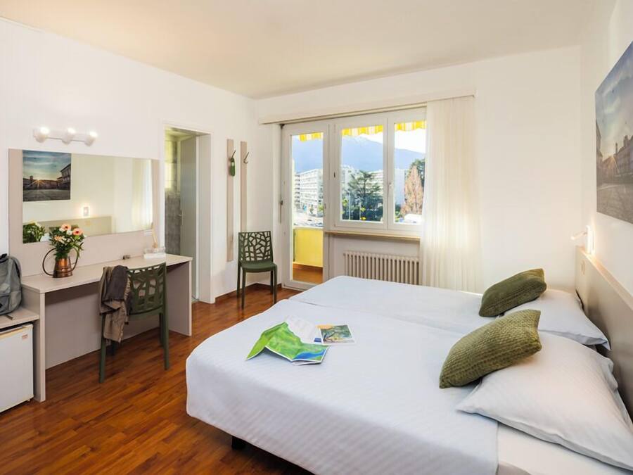 Habitación de hotel con cama, escritorio, silla y ventana con vistas a edificios y montañas.