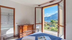 A bedroom with a bed, a wooden dresser, a glass shower door, and a balcony with a view of mountains and a lake. - Lenno Glamour (Tremezzina)