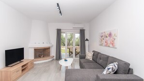 A modern living room with a flat-screen TV, a fireplace, a grey sofa, and a dining area visible through glass doors. - Nestled on the ground floor with direct garden access amidst olive trees (Psarou)