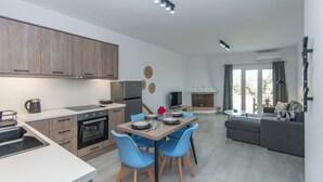 A modern kitchen with wooden cabinets, a dining area with blue chairs, a grey sofa, and a fireplace. - Nestled on the ground floor with direct garden access amidst olive trees (Psarou)