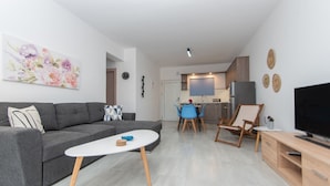 A modern living room with a grey sofa, a white coffee table, and a TV on a wooden stand. - Nestled on the ground floor with direct garden access amidst olive trees (Psarou)