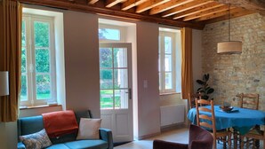 Ein gemütliches Wohnzimmer mit einem blauen Sofa, einem roten Sessel und einem Esstisch mit Stühlen. Große Fenster sorgen für natürliches Licht und Blick ins Grüne.