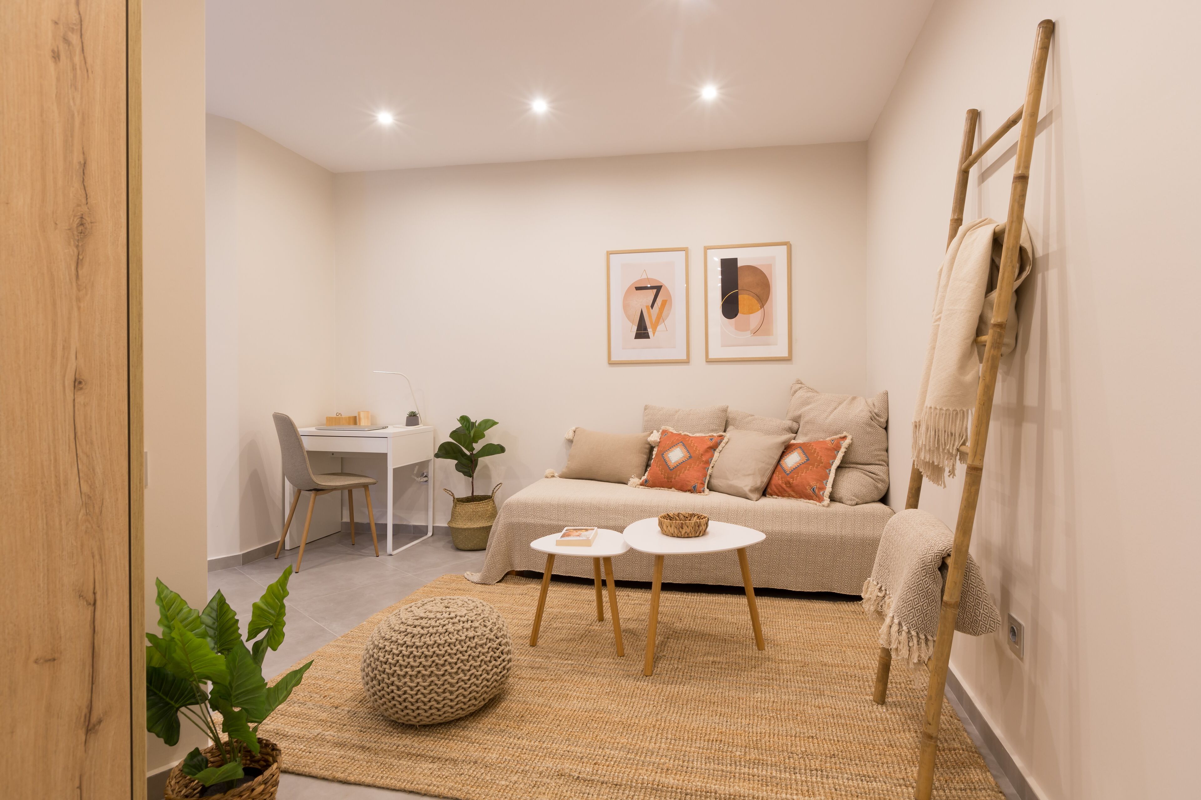 Een moderne woonkamer met een bank, twee bijzettafels, een klein bureau en een stoel. Twee ingelijste kunstwerken hangen aan de muur en een houten ladder leunt tegen de muur.