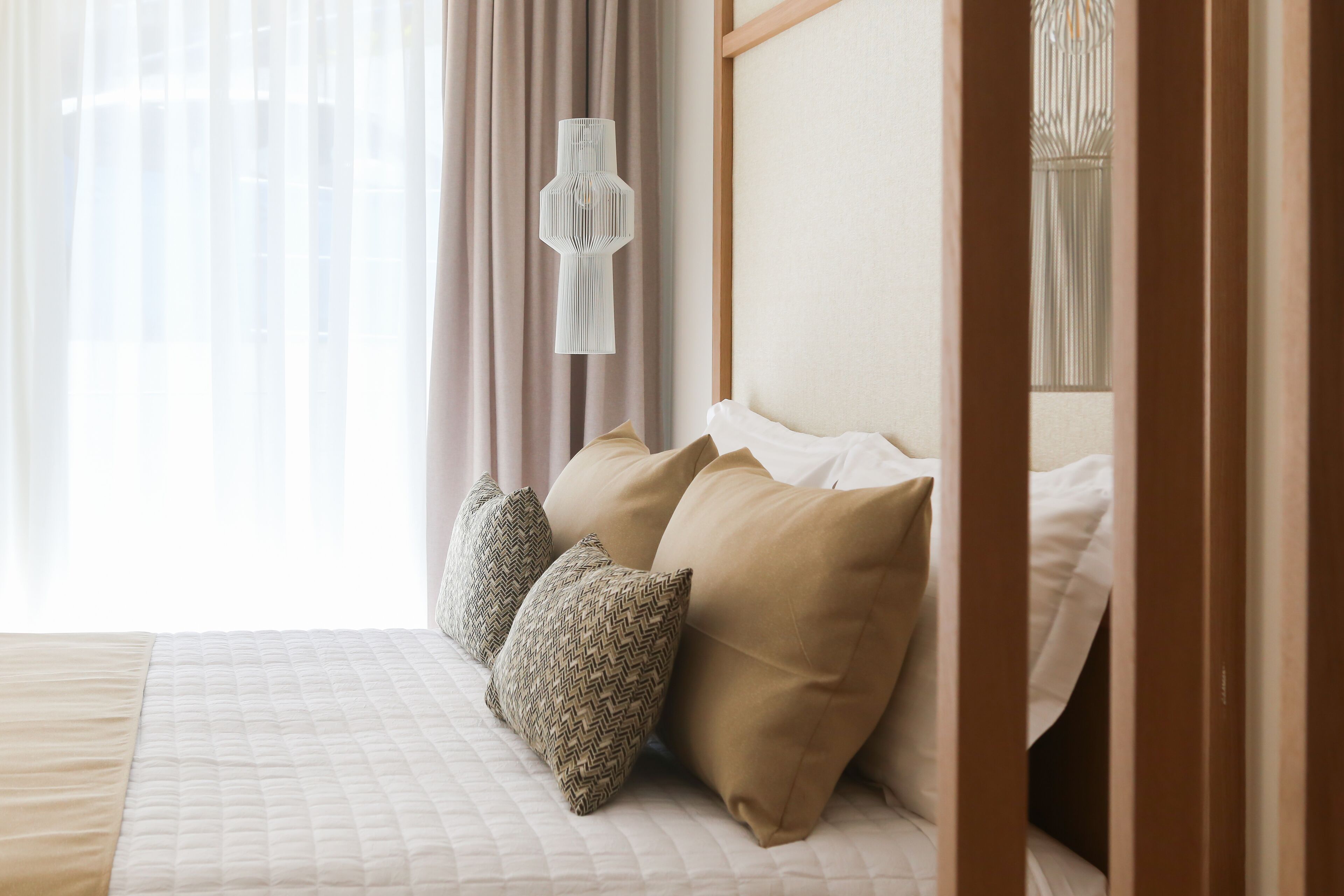 A neatly made bed with beige and patterned pillows, a hanging light fixture, and sheer curtains.
