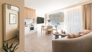 A modern living room with a grey sofa, a dining area with a round table and chairs, a kitchen with wooden cabinets and a TV, and a large window with sheer curtains. - Villa Iris (Mylopotamos)