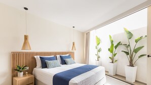 A modern bedroom with a wooden headboard, a large bed with a blue blanket, bedside tables with plants, and large windows with curtains. - Villa Iris (Mylopotamos)
