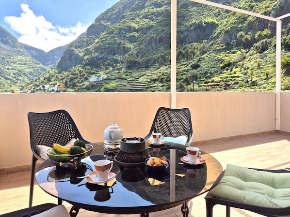Ein Balkon mit Tisch für zwei Personen, Stühlen und Blick auf grüne Hügel unter blauem Himmel.