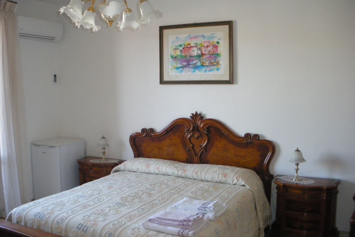 A bedroom with a wooden bed, a bedside table, a lamp, a wall-mounted air conditioner, and a framed painting.