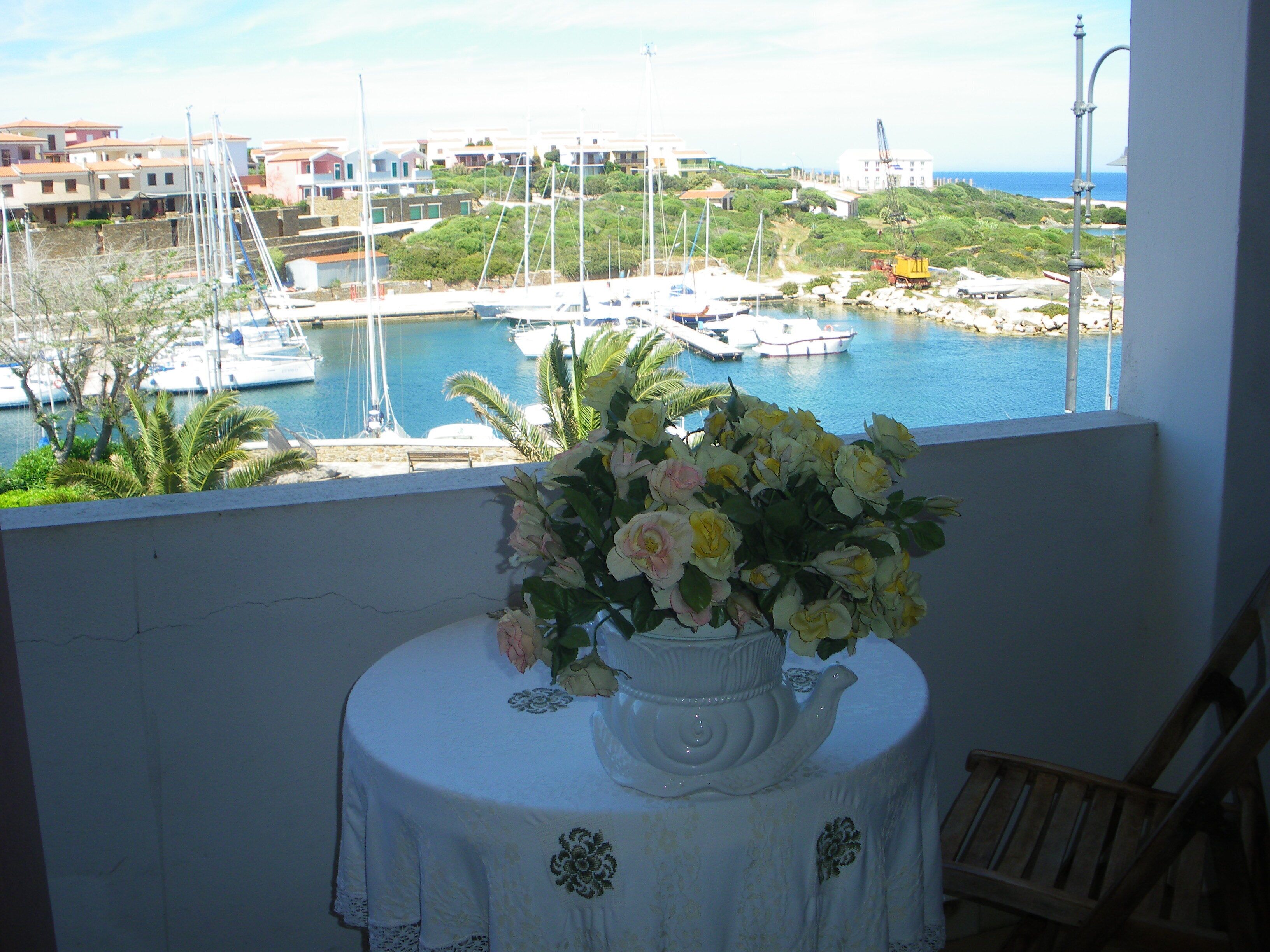 Un balcon avec une table sur laquelle est posé un vase de fleurs, donnant sur une marina avec des bateaux et des bâtiments.