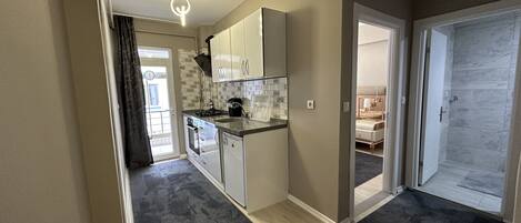 A modern kitchen with white cabinets, a black countertop, and a built-in oven. There is a door leading to a bedroom with a bed and a bathroom with a shower.