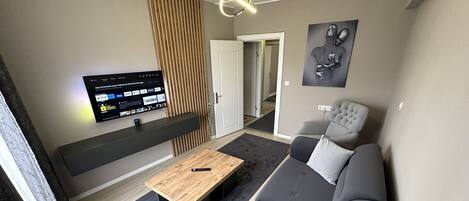 Una sala moderna con un sofá gris, una mesa de centro de madera, un televisor montado en la pared y una puerta que da a otra habitación.
