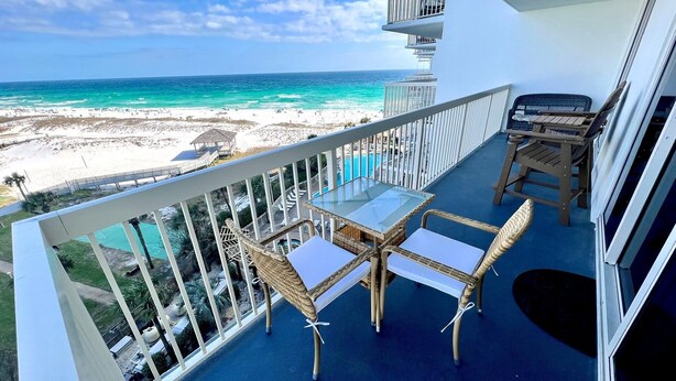 Een balkon met een glazen tafel en rieten stoelen, met uitzicht op een strand en een heldere blauwe oceaan.