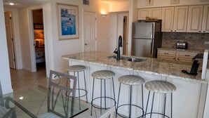 A kitchen with a granite countertop, stainless steel refrigerator, and bar stools. - Terrace At Pelican Beach 0507 3 Bedroom Condo (Destin)