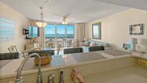 A spacious living room with a large window overlooking the beach, a ceiling fan, and a dining area with a view.