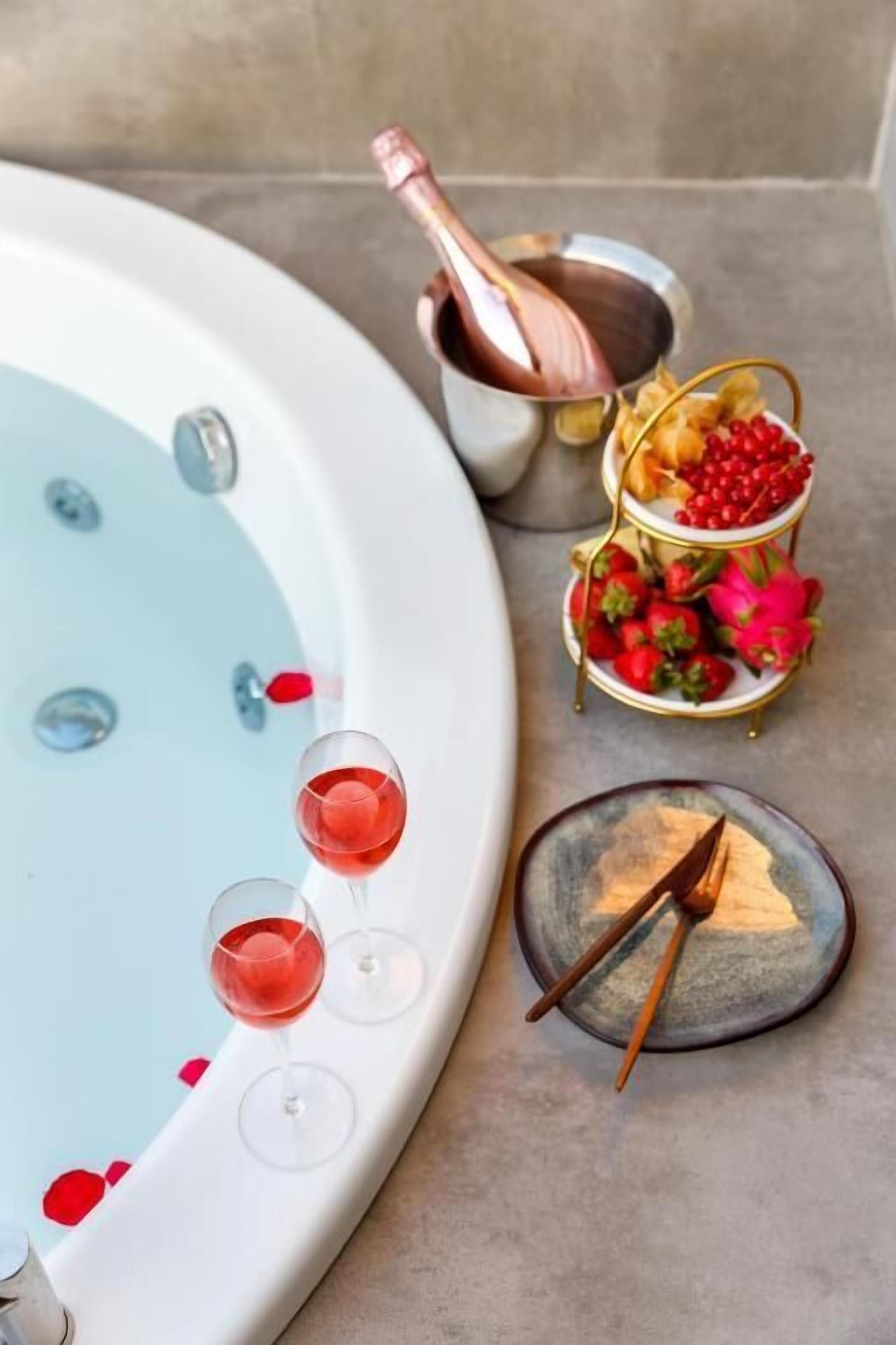 A bathtub with rose petals, a champagne bottle, and glasses of red wine.