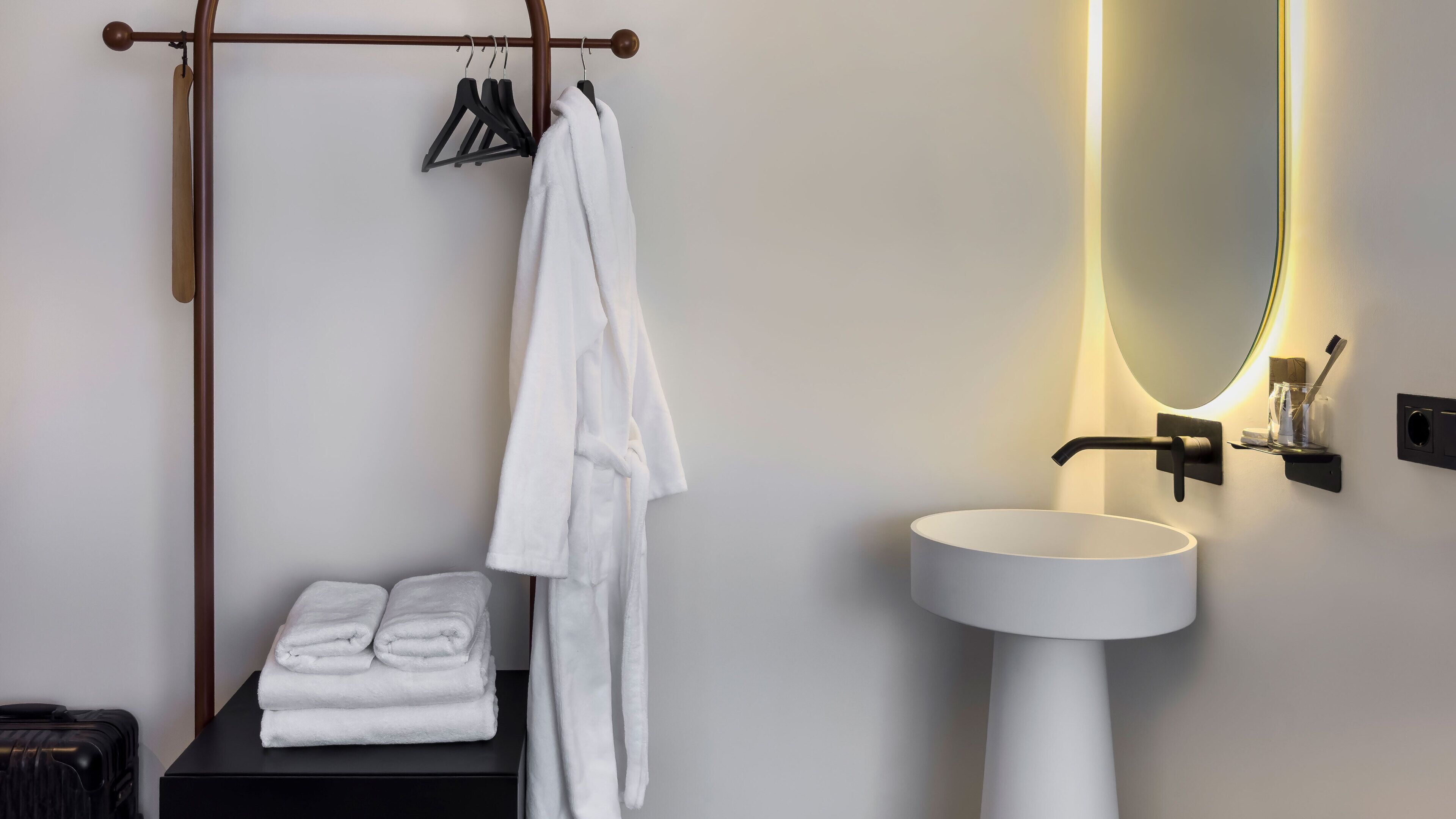 Un baño moderno con un lavamanos blanco, un espejo con iluminación integrada, un perchero y toallas dobladas cuidadosamente sobre una mesa negra.