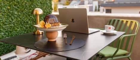 A table with a laptop, a cup, and a dessert on a terrace with a green hedge and chairs.