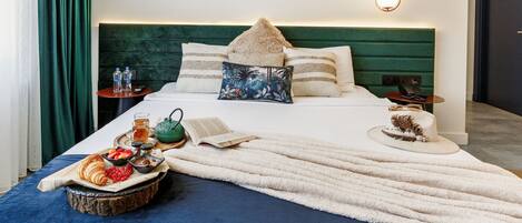 Una cama bien tendida con cabecera verde, una mesita de noche con lámpara y una bandeja con comida y bebidas.