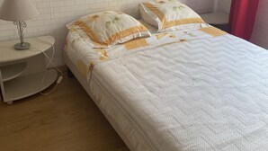 Een opgemaakt bed met een deken in wit en geel, een bedlamp en een witte bakstenen muur.