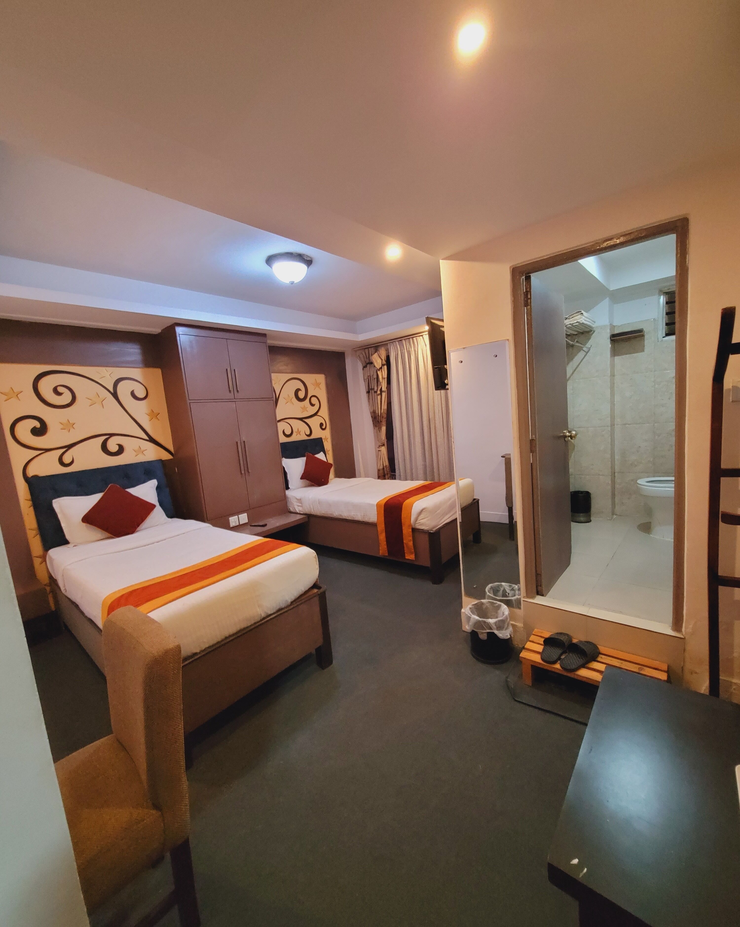 A hotel room with two single beds, a small table, a mirror, and a shower area visible through a glass door.