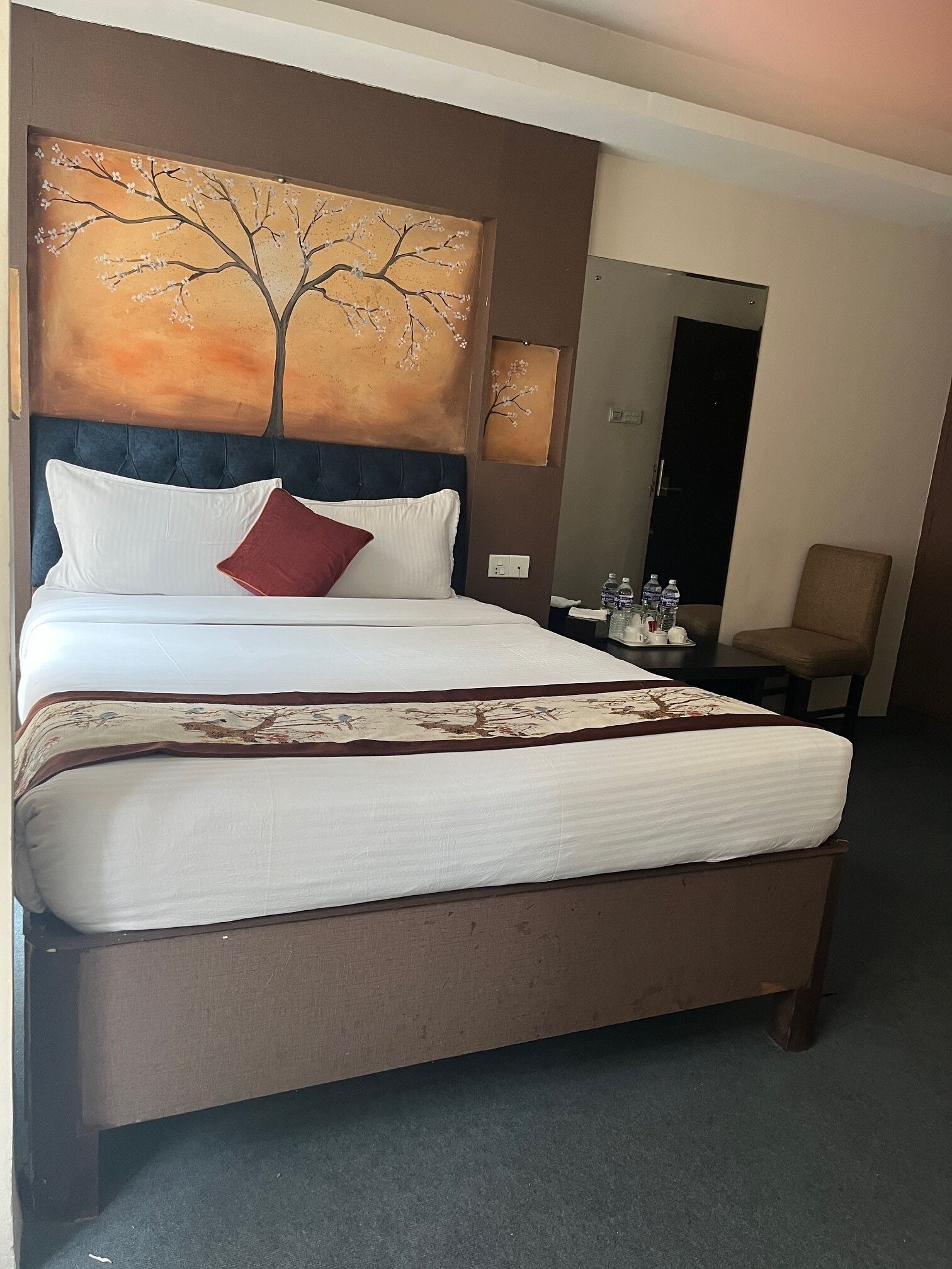A hotel room with a large bed, a headboard with a tree painting, a small table with water bottles, and a chair.