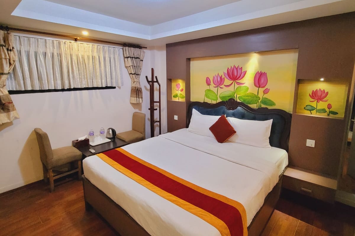 Quarto de hotel com cama grande, piso de madeira, escrivaninha com cadeira, janela com cortinas e luminária no teto.